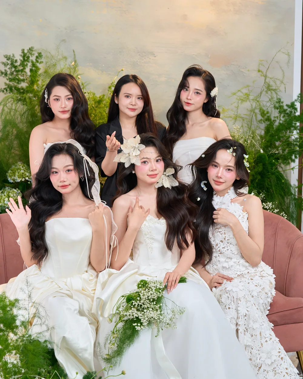 Nhóm cô dâu do AMI Makeup trang điểm — váy cưới trắng, hoa cài tóc, bên ghế hồng giữa nền hoa lá